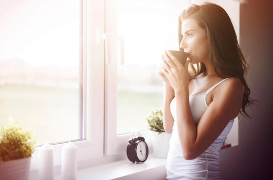 Woman sipping from a mug by a sunny window in the morning, alarm clock on the sill — supporting sleep and gut health with a morning routine