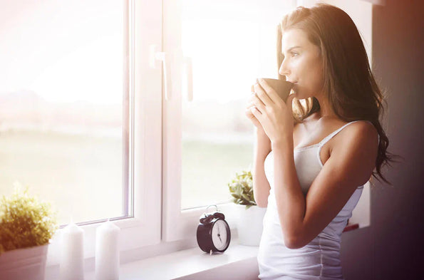 Woman sipping from a mug by a sunny window in the morning, alarm clock on the sill — supporting sleep and gut health with a morning routine